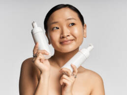 A young Asian woman smiles joyfully holding two skincare products against a light background.